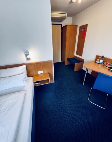 Einfaches Hotelzimmer mit Bett, Schreibtisch und blauem Teppich.