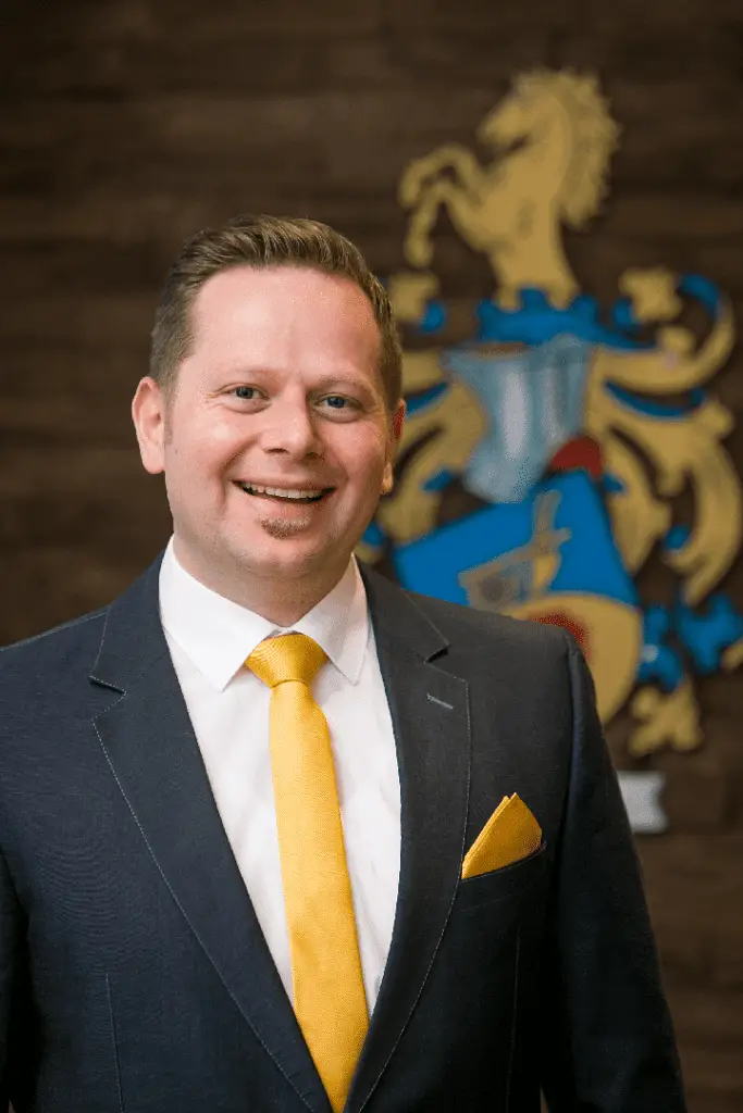 Lächelnder Mann im Anzug mit Krawatte und Wappen.